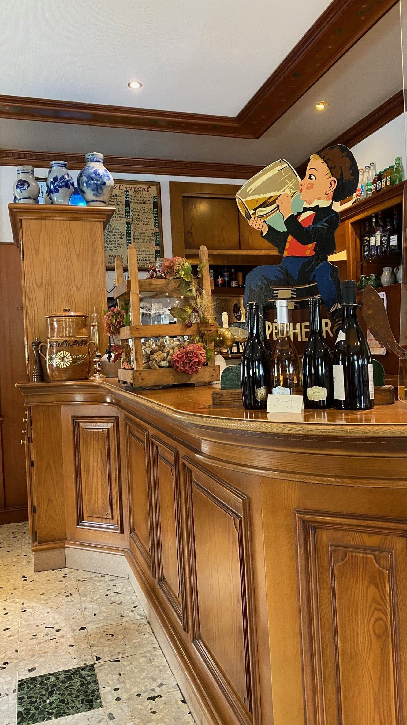 Comptoir en bois courbé d’un restaurant alsacien avec bouteilles de vin, poteries décoratives et figure vintage, créant une ambiance chaleureuse et traditionnelle.