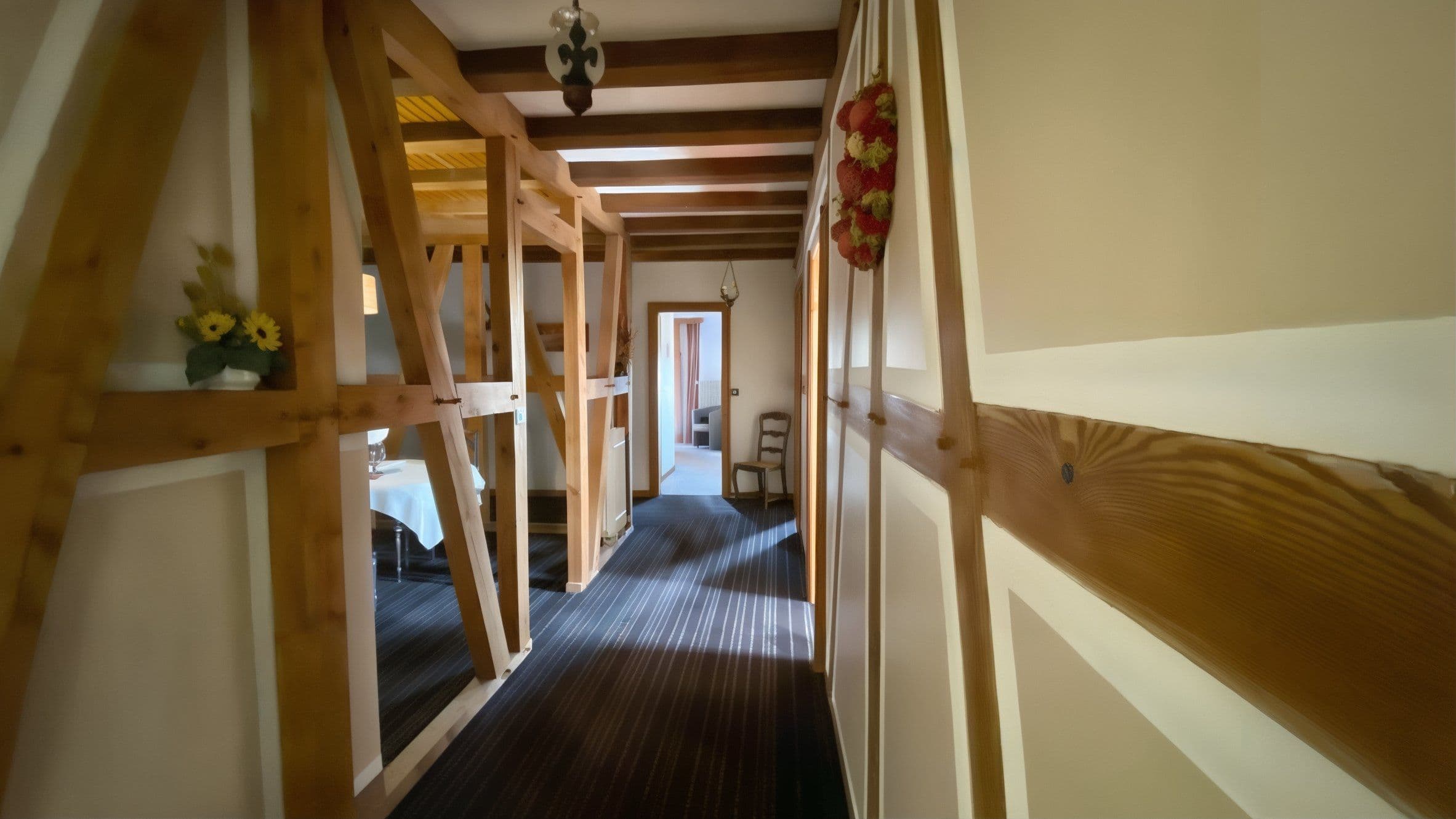 Couloir étroit aux poutres apparentes de style alsacien menant vers une chambre, avec moquette rayée et petites décorations traditionnelles.