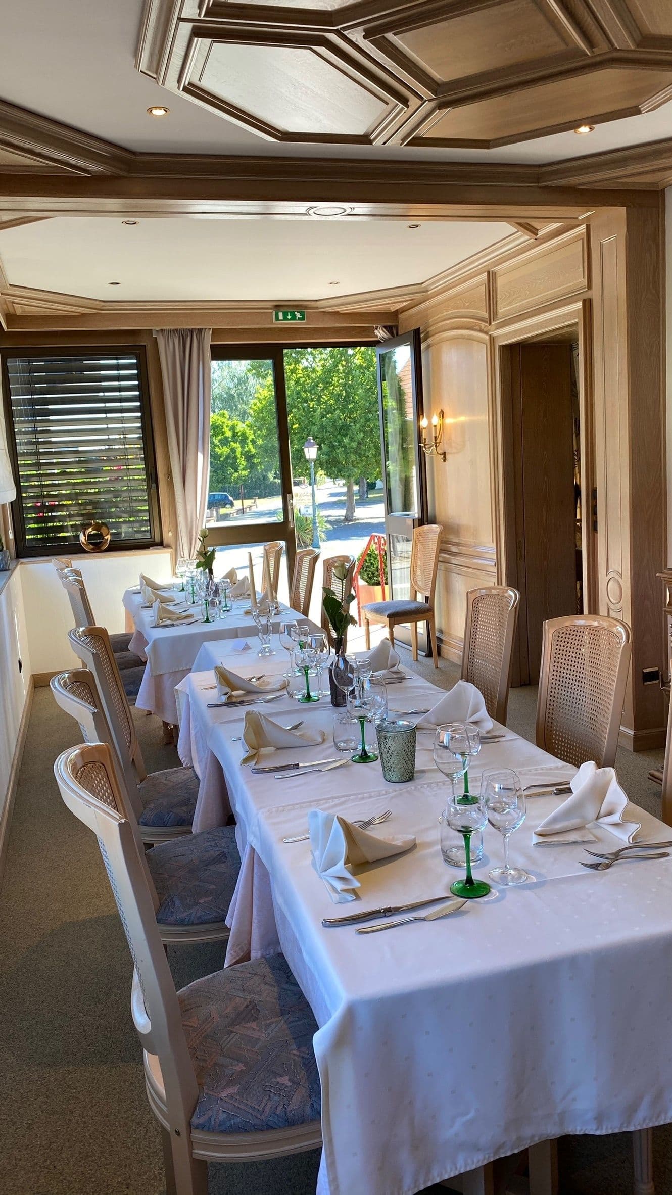 Salle à manger privée traditionnelle à pans de bois, table longue dressée pour un repas formel avec nappes blanches, verres à tige verts et couverts polis, baignée de lumière naturelle et ouverte sur le jardin.