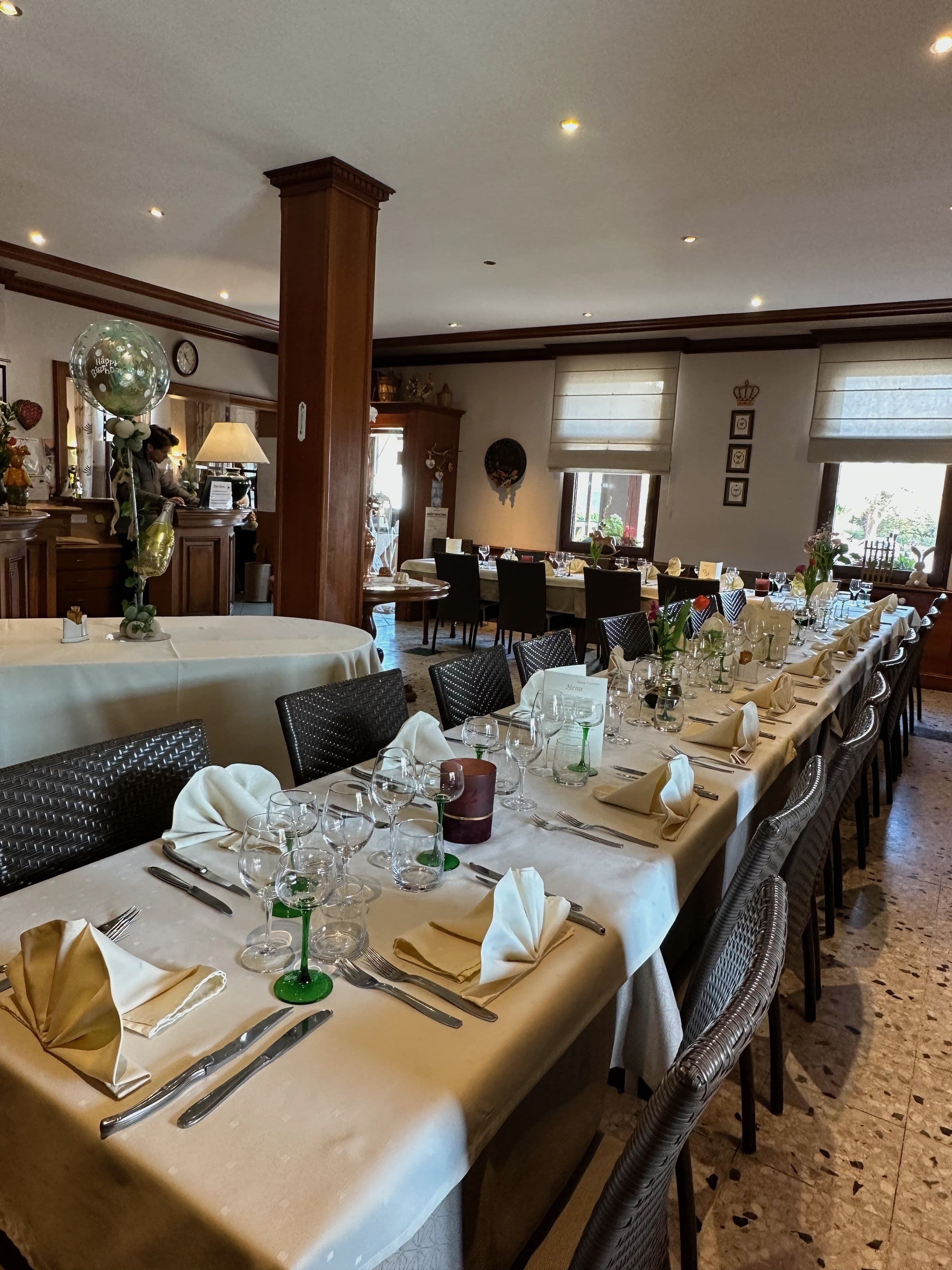 Salle à manger de l’hôtel prête pour un banquet : longues tables nappées, serviettes pliées, verres et couverts disposés dans une ambiance chaleureuse et boisée.