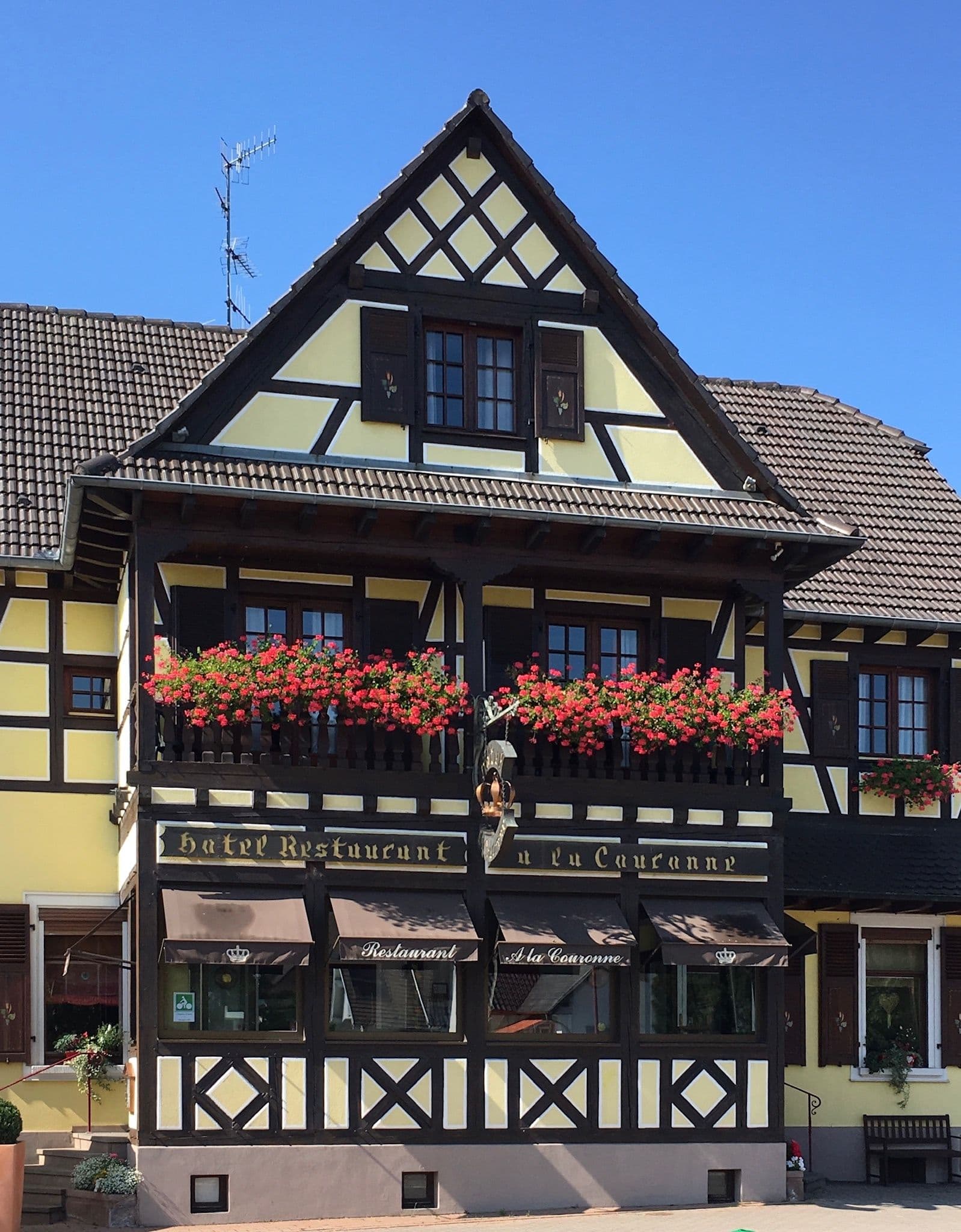 Façade à colombages d’un hôtel-restaurant alsacien jaune et bois foncé avec balcon garni de géraniums rouges, enseigne sculptée et auvents du restaurant au rez-de-chaussée.