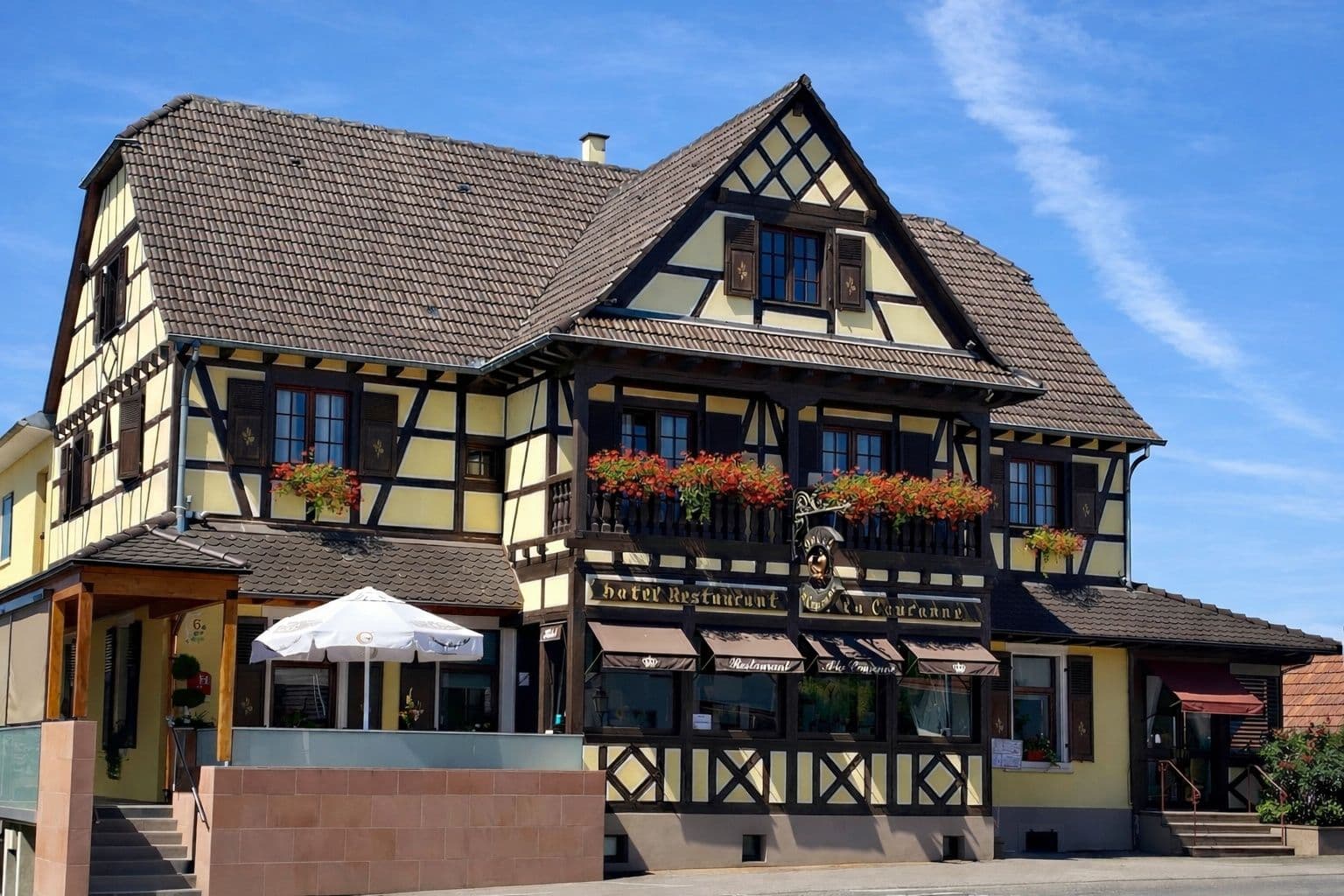 Façade à colombages de l'Hôtel-Restaurant À La Couronne à Kilstett, avec balcons fleuris, terrasse ombragée et enseigne traditionnelle sous un ciel bleu.