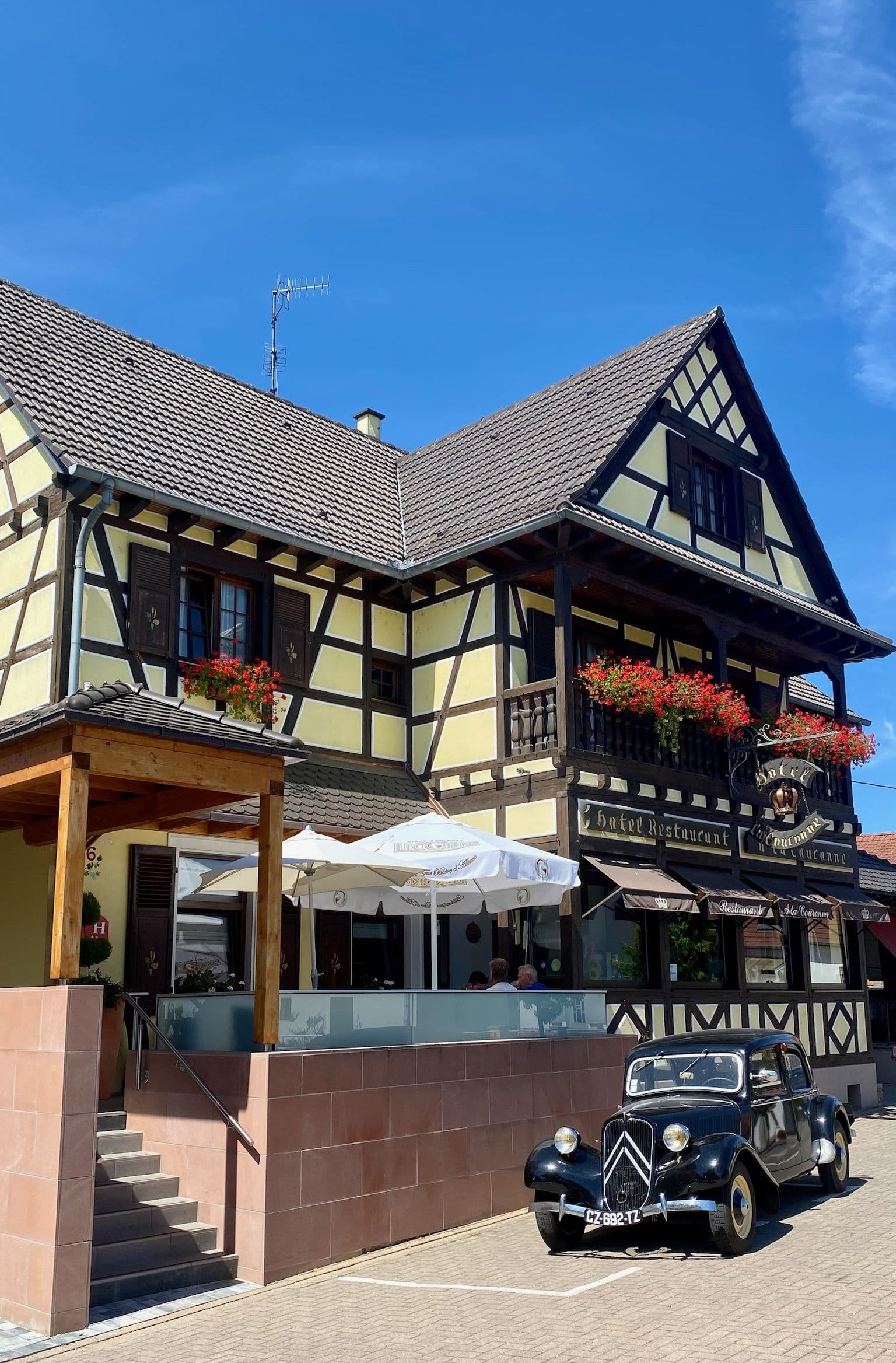Façade à colombages d’un hôtel-restaurant alsacien sous un ciel bleu, balcon et fenêtres fleuris, terrasse couverte avec parasols et une voiture Citroën ancienne garée devant.