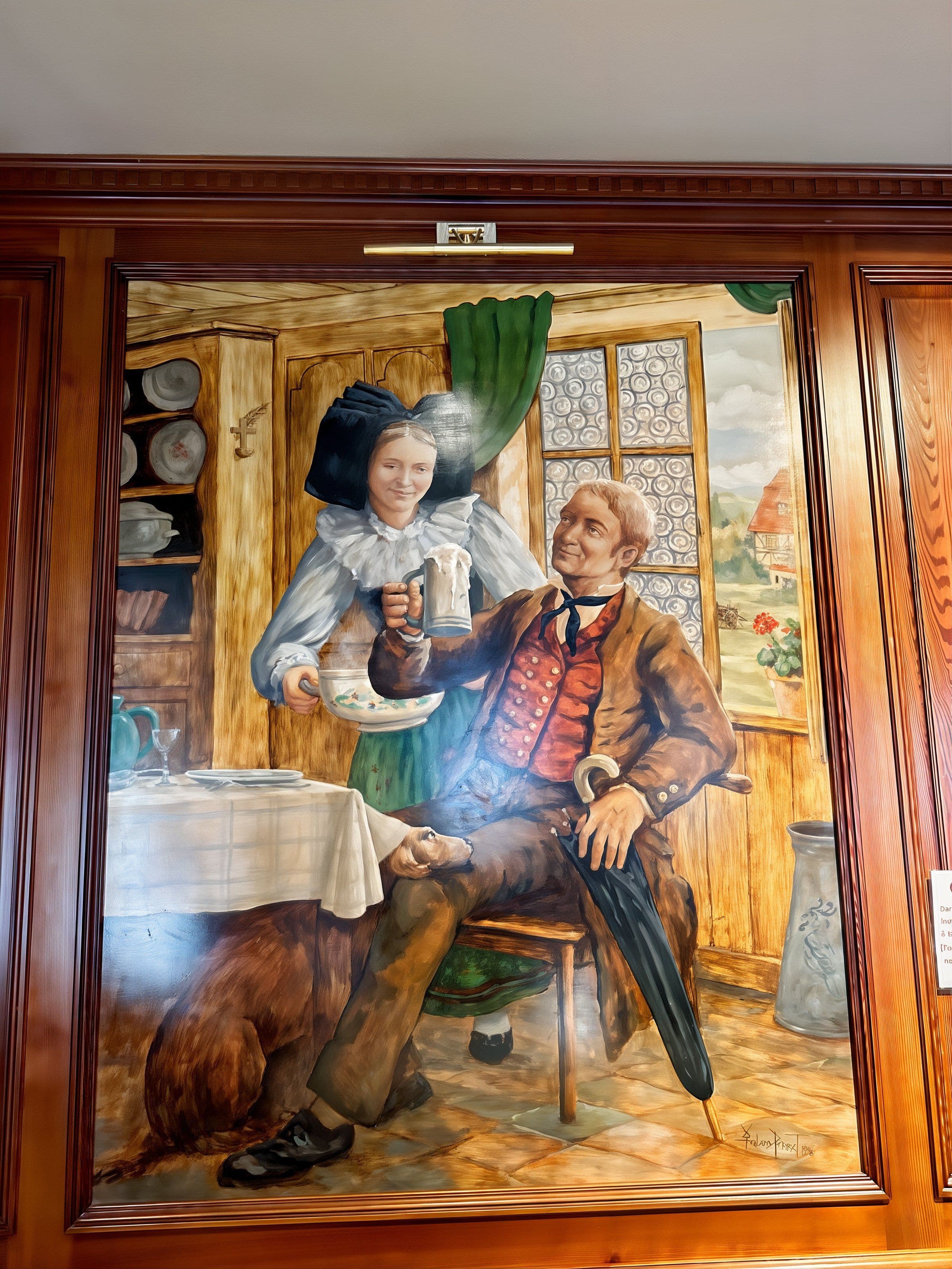 Grande fresque murale aux tons chauds représentant une scène alsacienne traditionnelle : un homme assis lève sa chope tandis qu'une femme en costume régional le sert, avec un chien à ses pieds et une fenêtre ouvrant sur une maison colombage. L'œuvre, encadrée de boiseries polies, orne la salle du restaurant et renforce l'ambiance locale et conviviale.