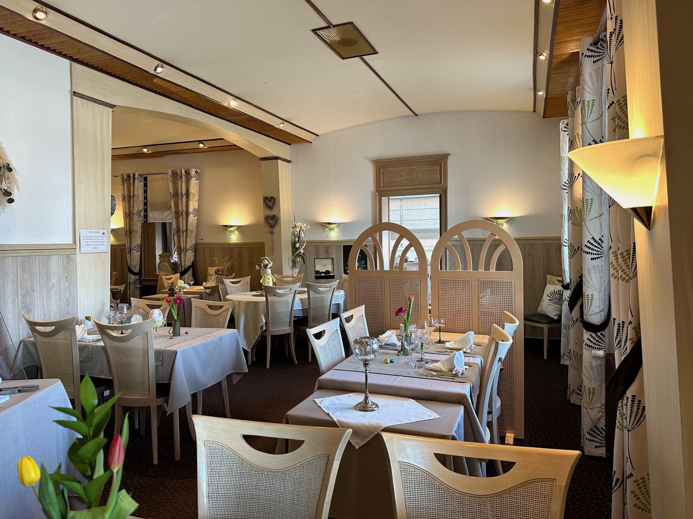 Salle à manger traditionnelle alsacienne lumineuse avec tables nappées, chaises en bois clair, écrans de séparation et décor floral, créant une ambiance chaleureuse et intime.