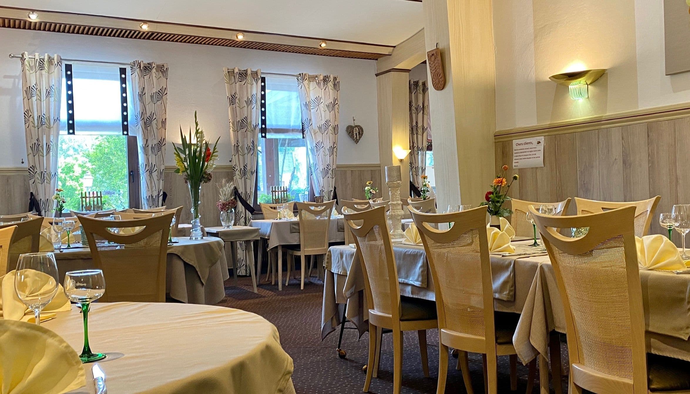 Salle à manger traditionnelle de l’hôtel-restaurant avec tables dressées, chaises en bois clair, rideaux à motif et bouquets de fleurs, éclairage doux créant une atmosphère chaleureuse d’Alsace.