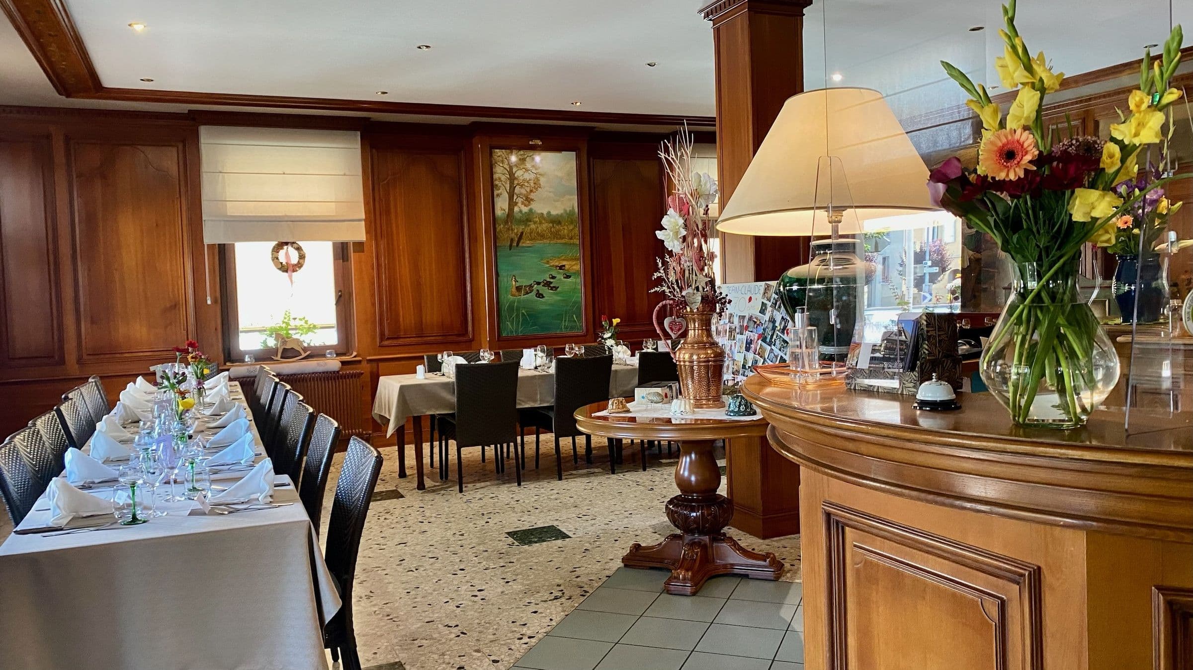 Salle à manger alsacienne chaleureuse avec boiseries, longues tables dressées pour le service et comptoir en bois orné de fleurs et d'une lampe.