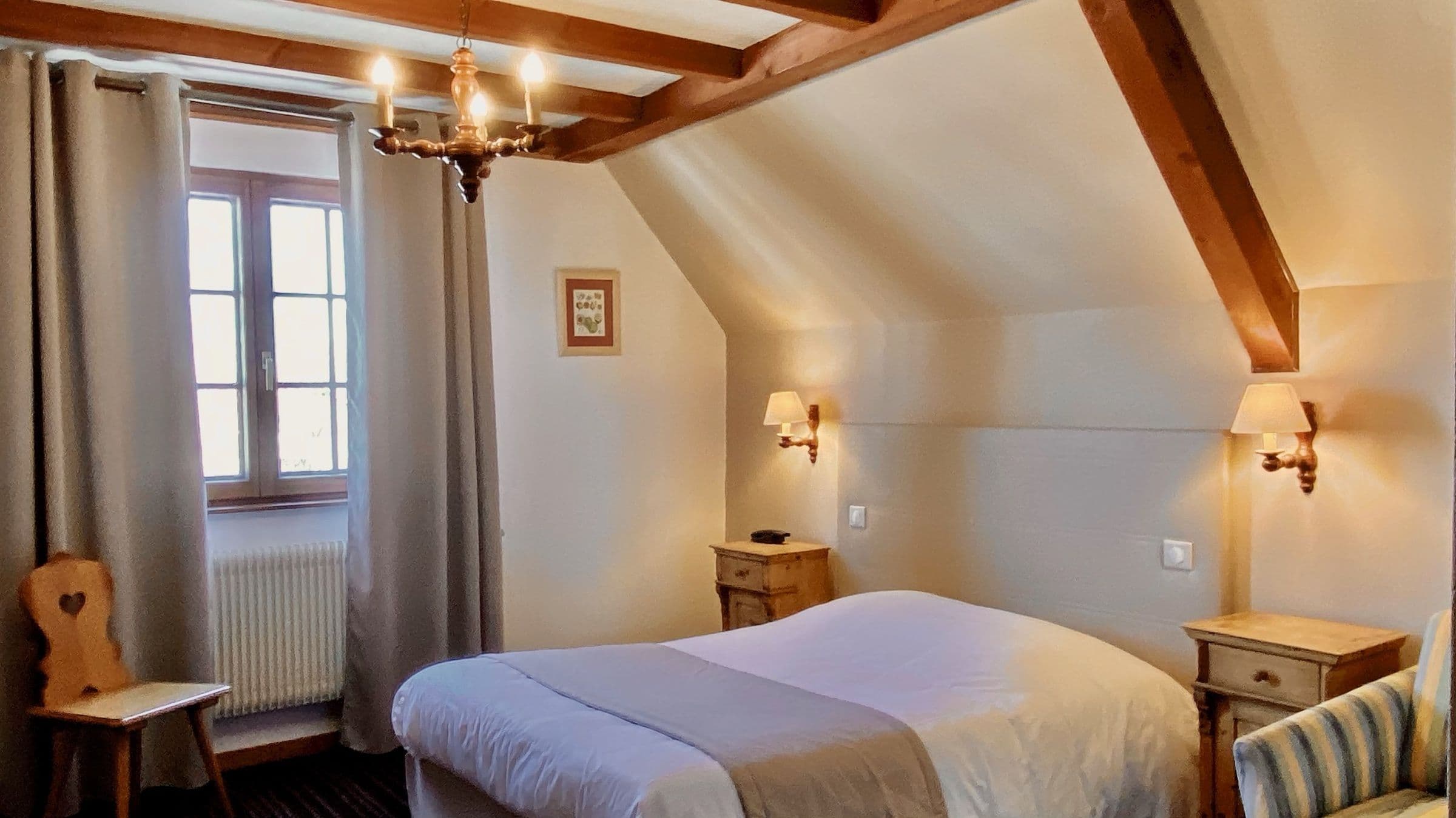Chambre double mansardée aux poutres apparentes, lit bien fait, chevets en bois et lampes murales chaudes, fenêtre avec rideaux et chaise traditionnelle.