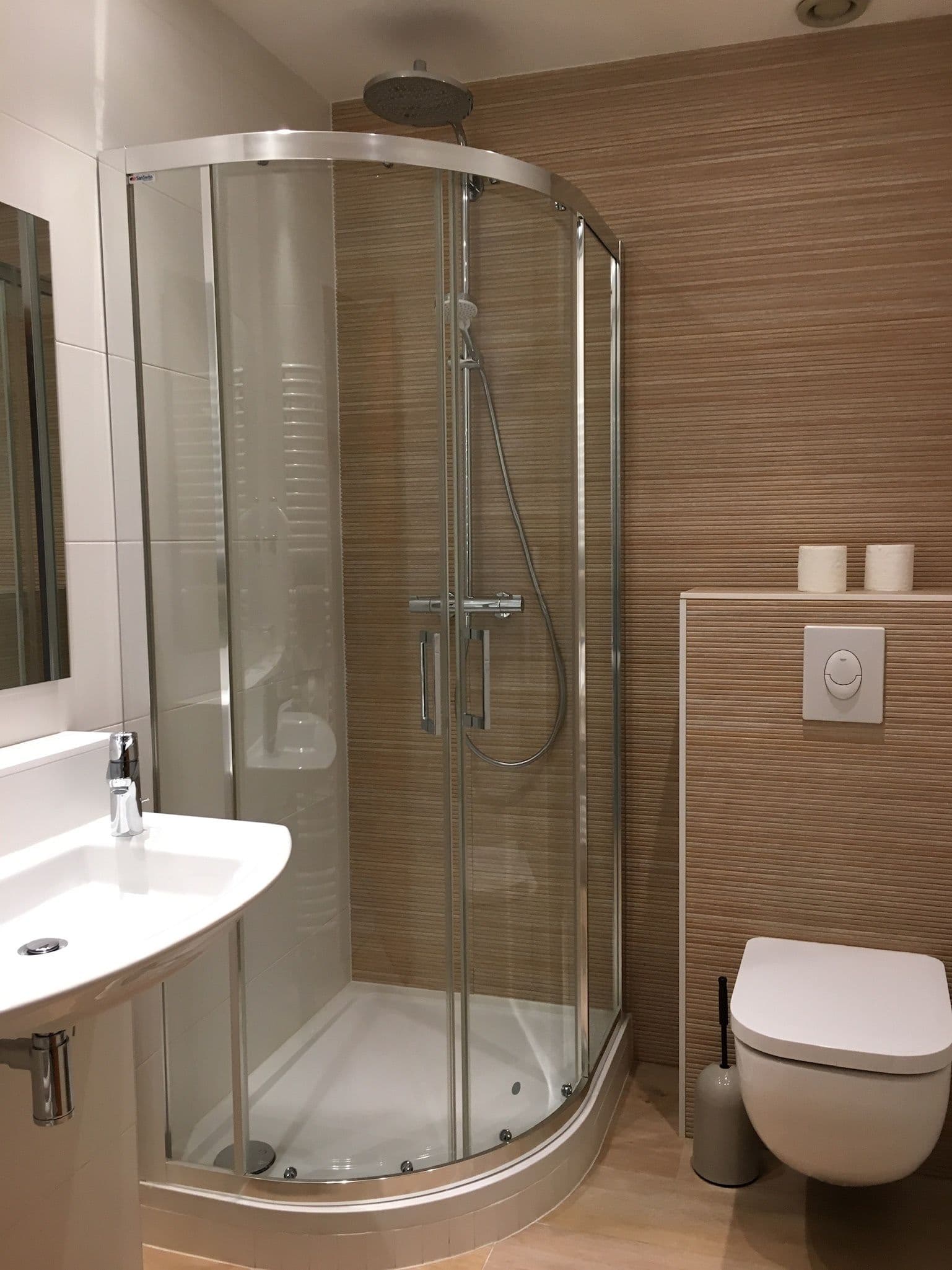 Salle de bain privative moderne avec cabine de douche d'angle en verre, lavabo blanc et toilettes suspendues sur fond de carrelage effet bois.