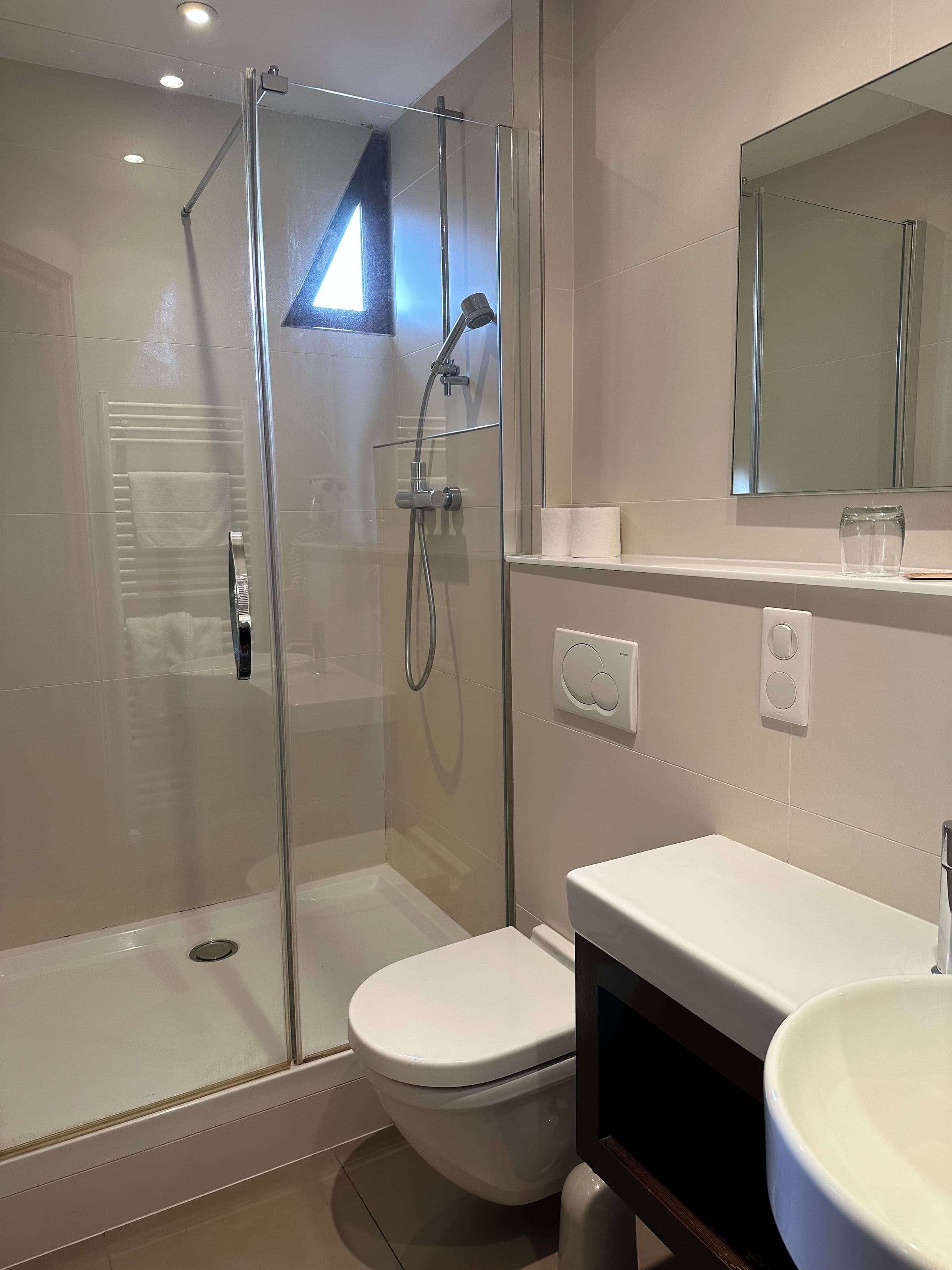 Salle de bains d’une chambre avec cabine de douche vitrée, douchette à main, toilette suspendue et petit lavabo sur meuble, ambiance épurée et moderne.