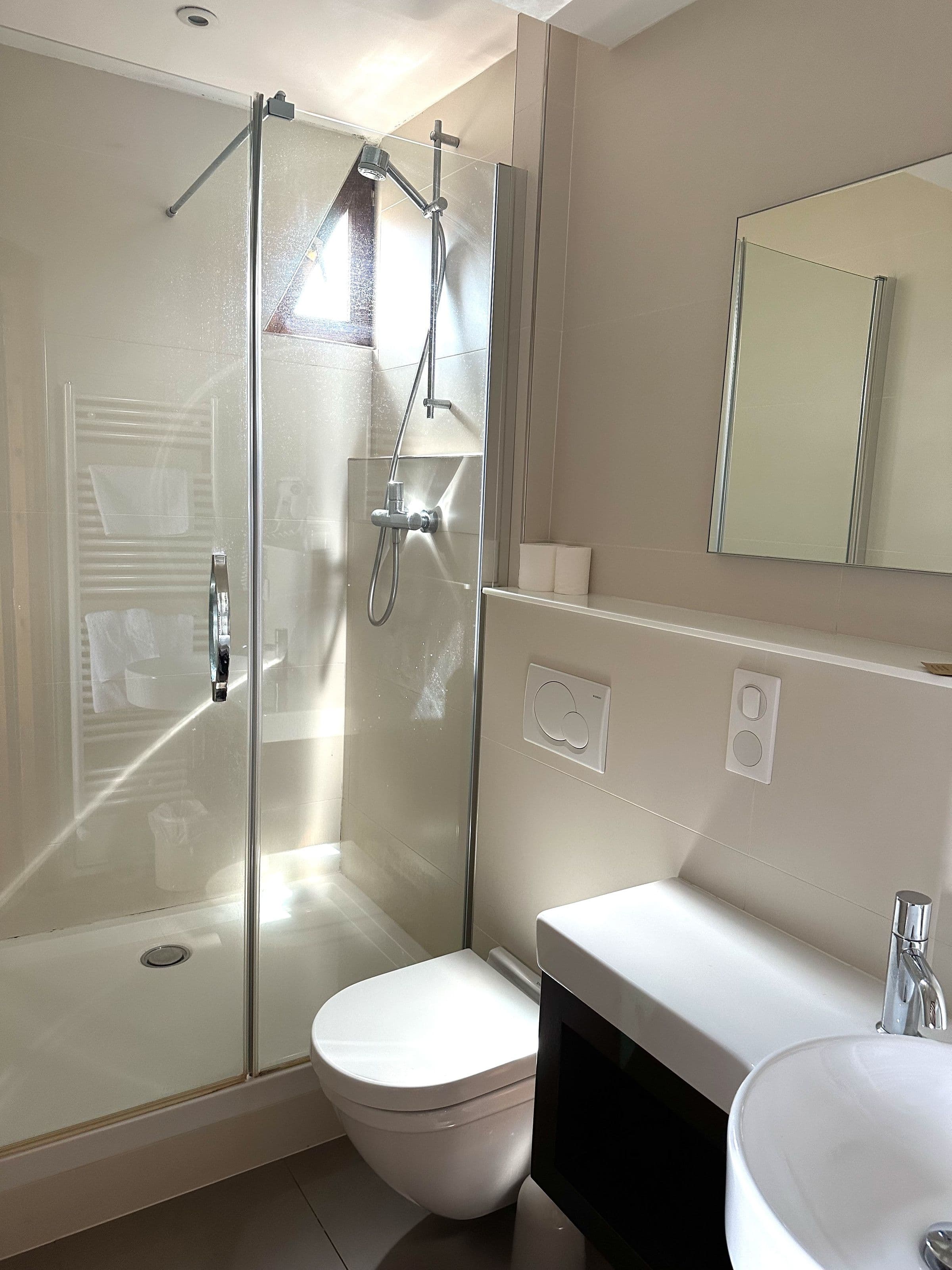 Salle de bains d’une chambre avec douche vitrée, robinetterie chromée, WC suspendu et lavabo compact, éclairée par une petite fenêtre triangulaire.