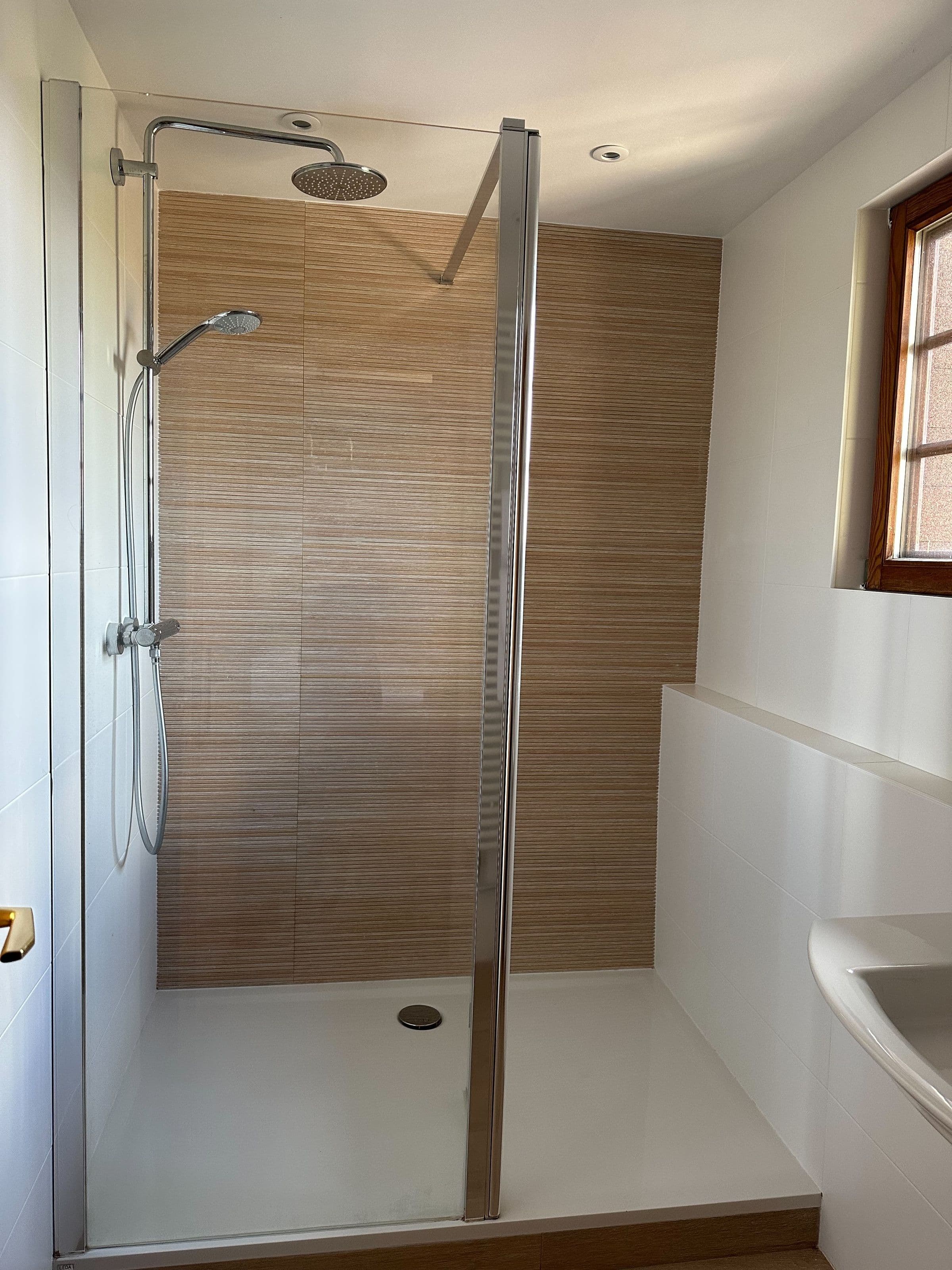 Douche à l'italienne moderne et lumineuse avec paroi vitrée, pommeau de pluie et carrelage effet bois, lavabo blanc et fenêtre encastrée.