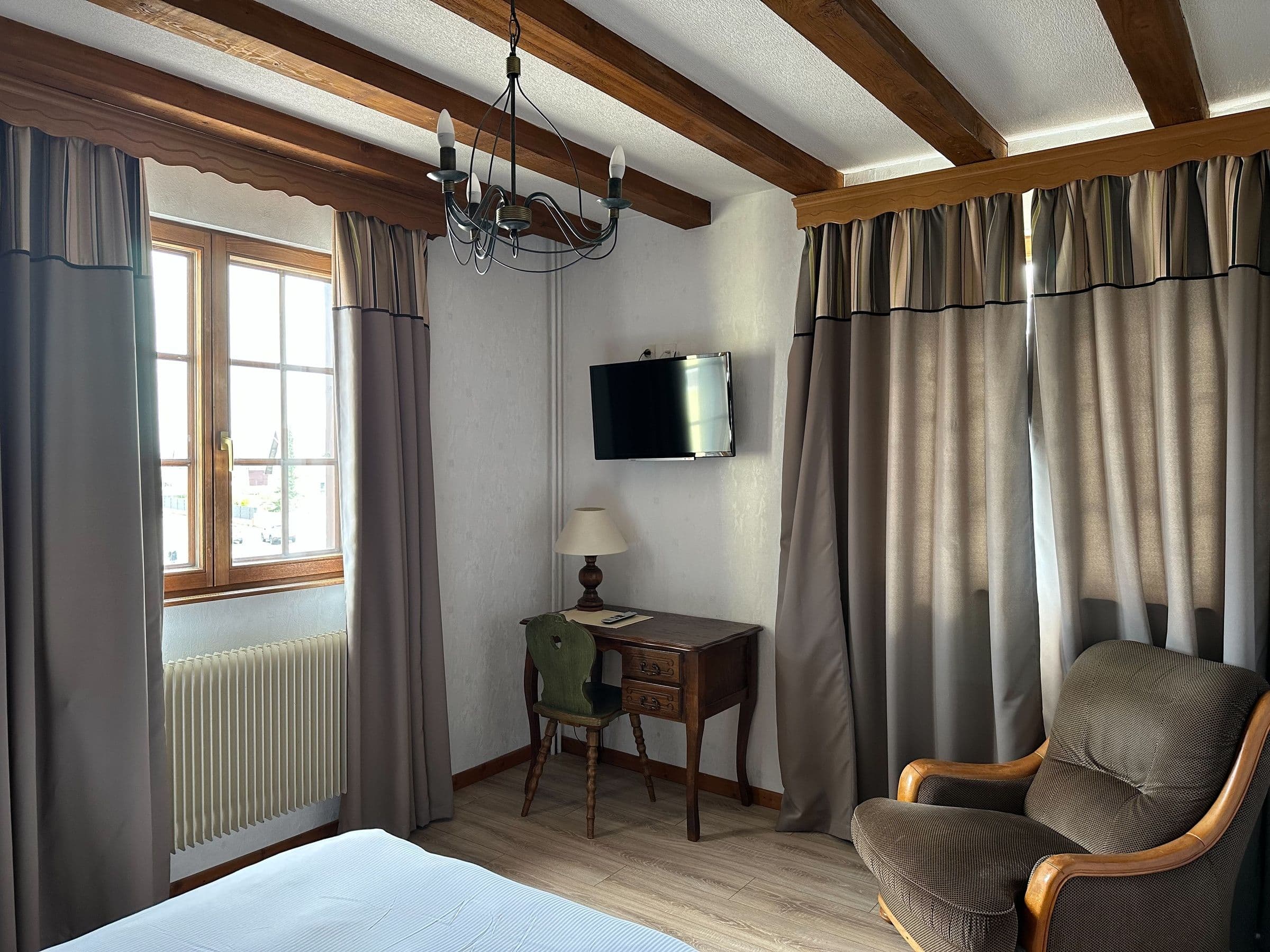 Chambre d'hôtel traditionnelle éclairée par la lumière du jour, avec poutres apparentes, fauteuil rembourré, petit bureau ancien et télévision murale, rideaux épais encadrant une fenêtre en bois.