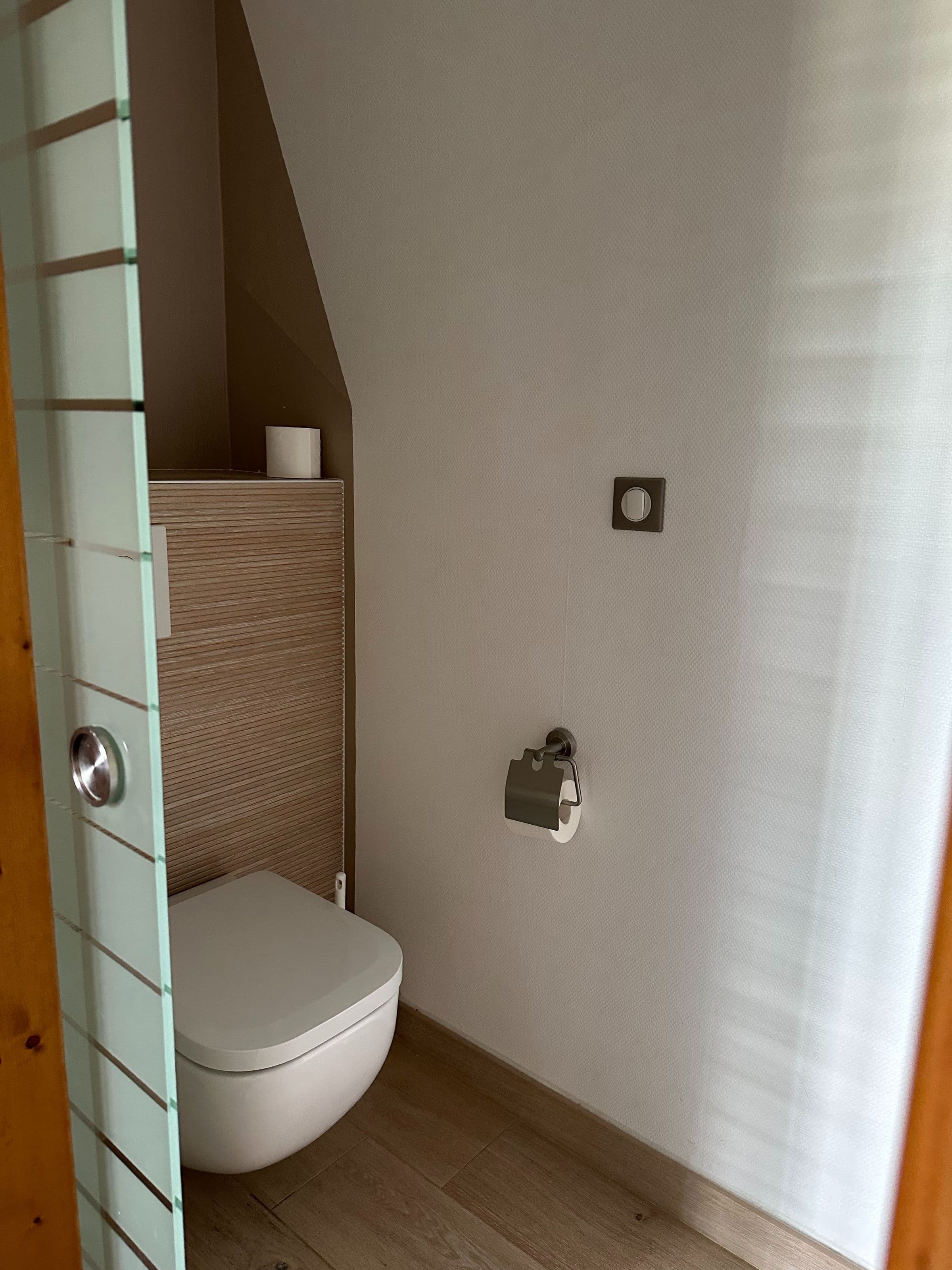 Petite toilette d’une chambre d’hôtel avec WC suspendu, placage bois clair, porte en verre dépoli et porte-rouleau de papier hygiénique métallique.