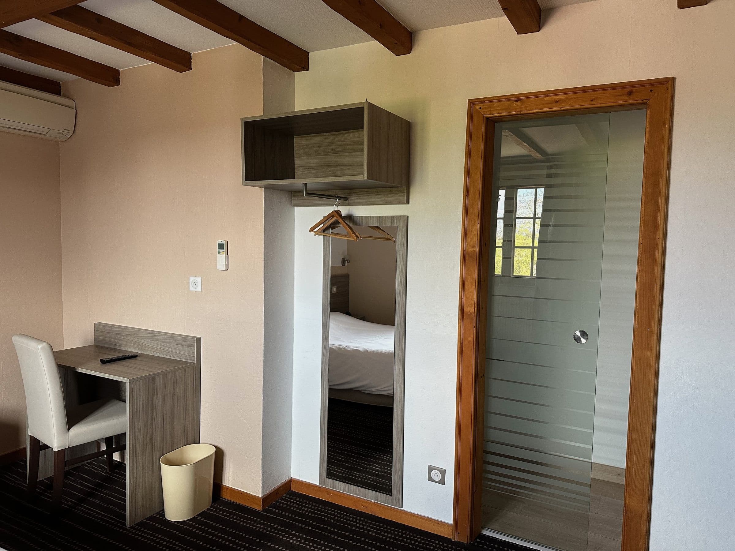 Chambre double lumineuse avec bureau en bois et chaise, étagère ouverte avec cintres et miroir plein pied reflétant le lit; porte de salle de bain en verre sablé encadrée de bois et poutres apparentes au plafond.
