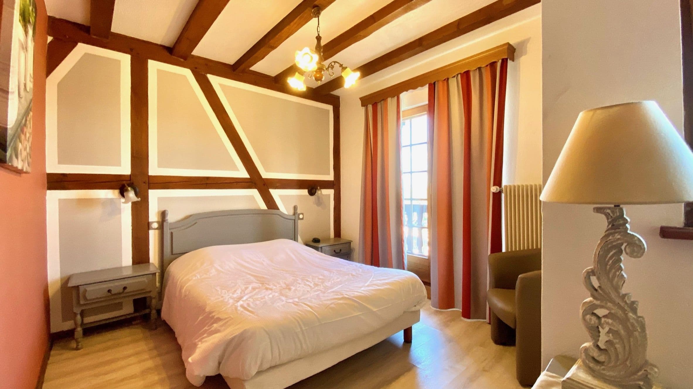 Chambre double chaleureuse dans une maison à colombages alsacienne, avec poutres apparentes, lit double, tables de chevet et porte-fenêtre donnant sur un petit balcon.