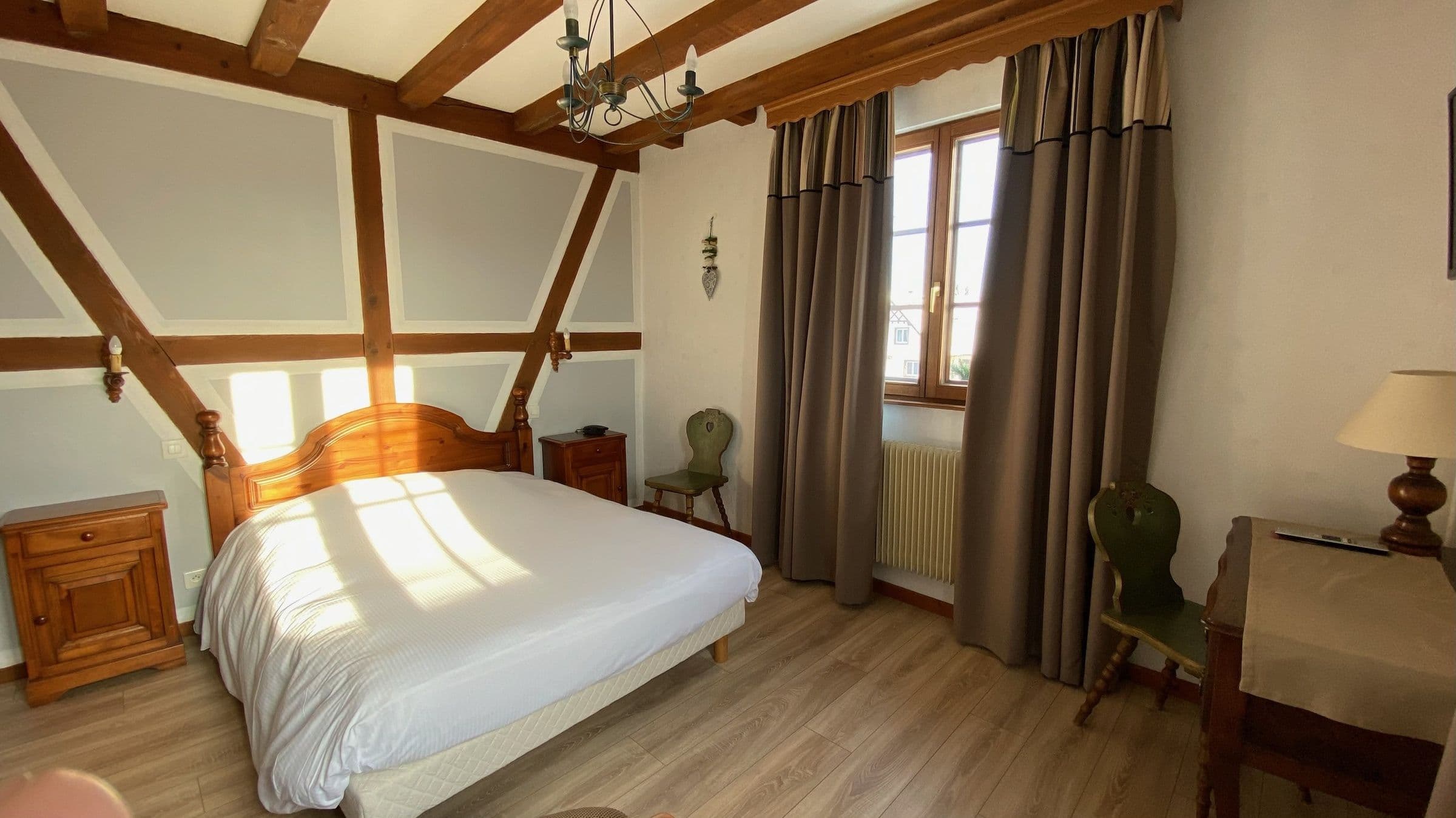 Chambre double ensoleillée avec poutres apparentes et mobilier en bois traditionnel, lit double, chevets, petit bureau et chaises décoratives près d'une fenêtre aux rideaux épais.