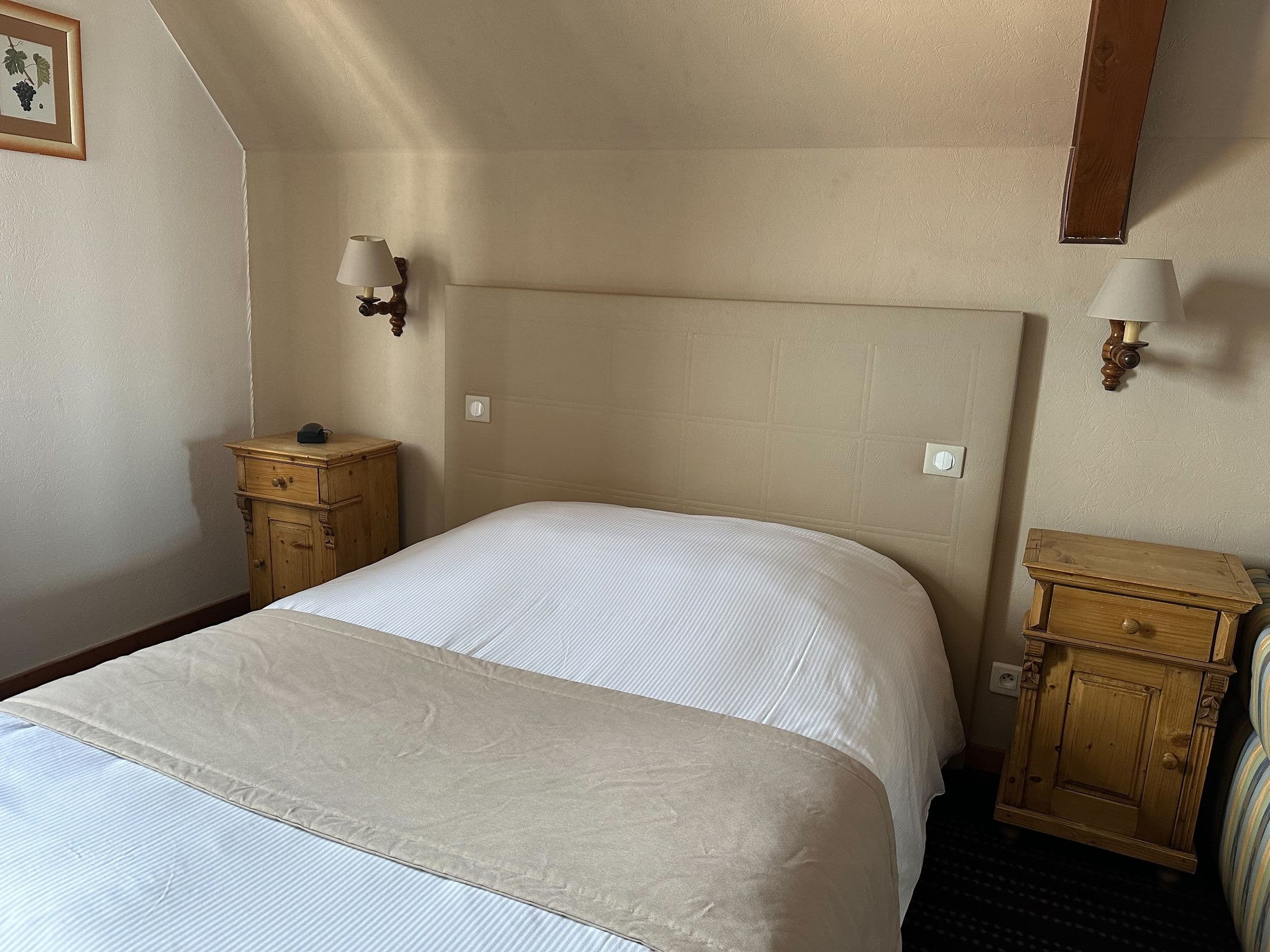 Chambre double accueillante avec tête de lit rembourrée, draps blancs et couvert beige, poutre apparente et chevets en pin pour une ambiance alsacienne traditionnelle.