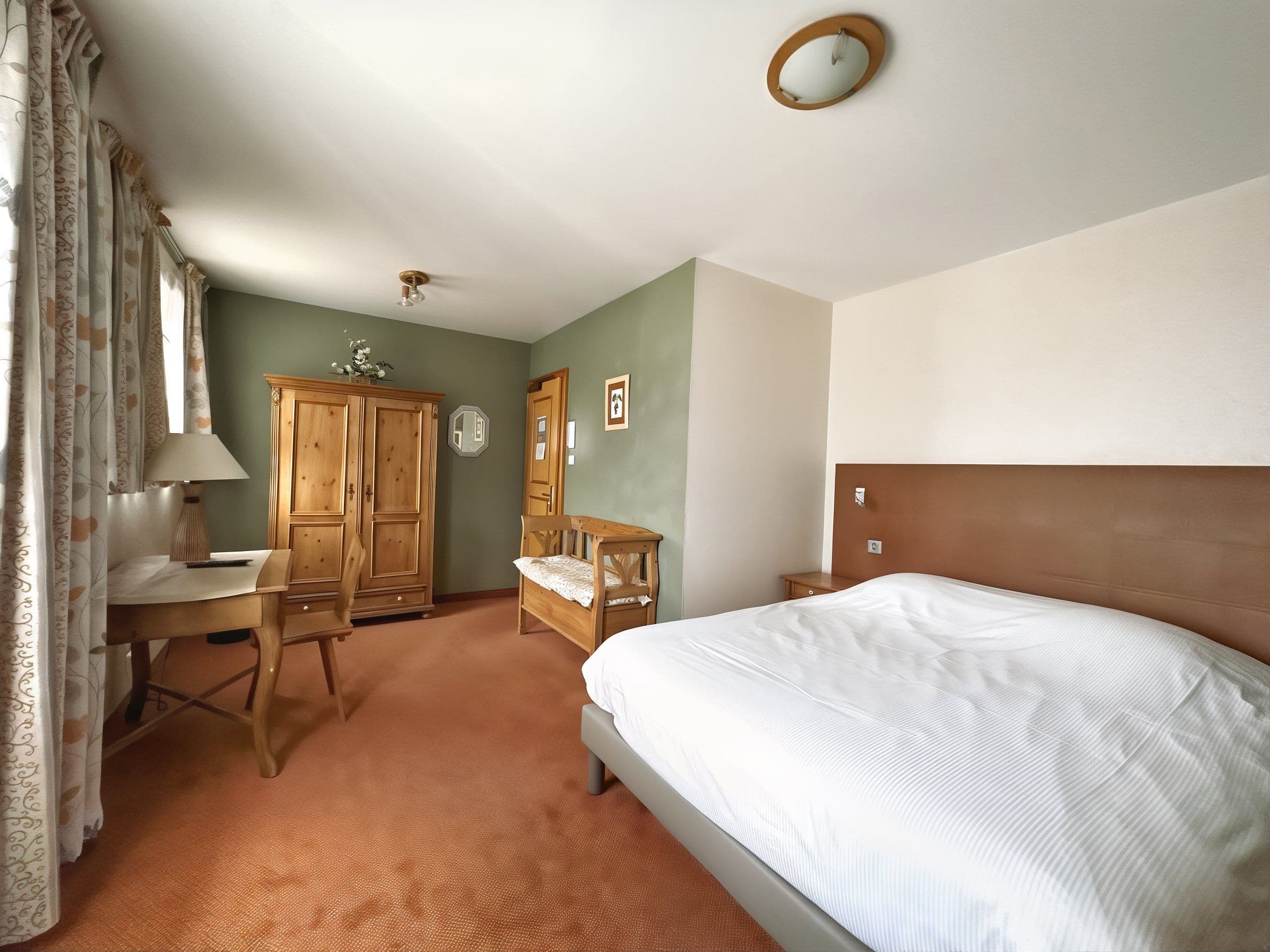 Chambre double chaleureuse au style traditionnel alsacien avec un grand lit en linge blanc, une armoire et un bureau en bois, un mur d'accent vert et des rideaux à motifs.