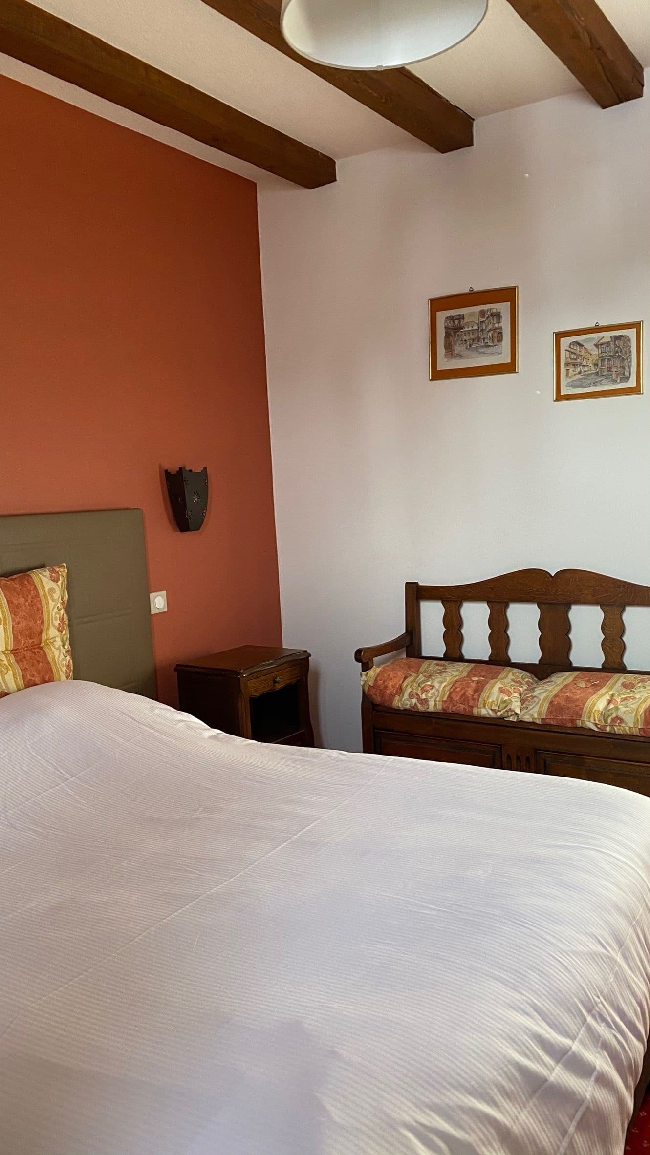 Chambre chaleureuse à l’alsacienne avec poutres apparentes, lit double en draps blancs, mur d’accent terracotta et banc en bois sculpté sous des estampes encadrées.