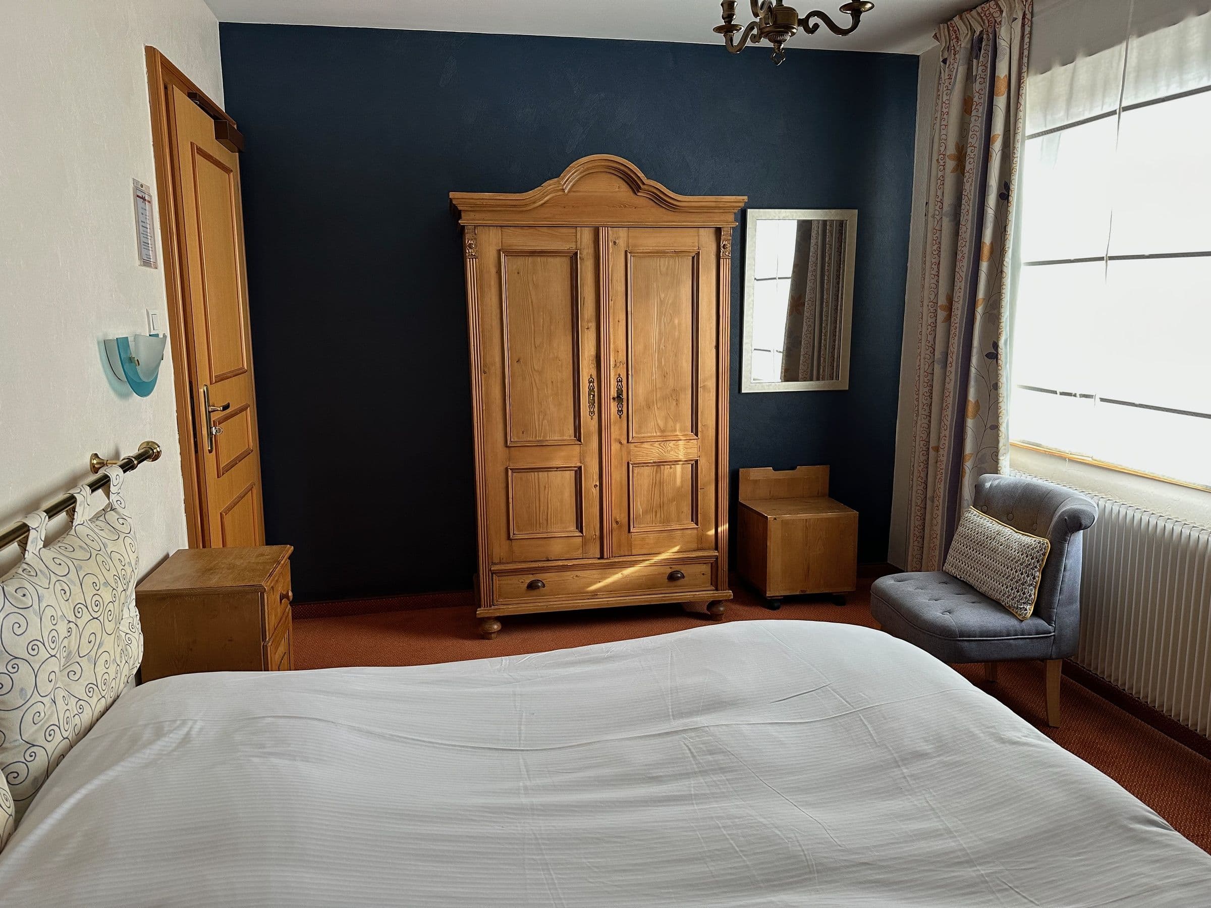 Chambre double confortable avec une grande armoire en bois sculpté, linge blanc, un fauteuil rembourré près de la fenêtre et un mur d'accent bleu, décor traditionnel alsacien.