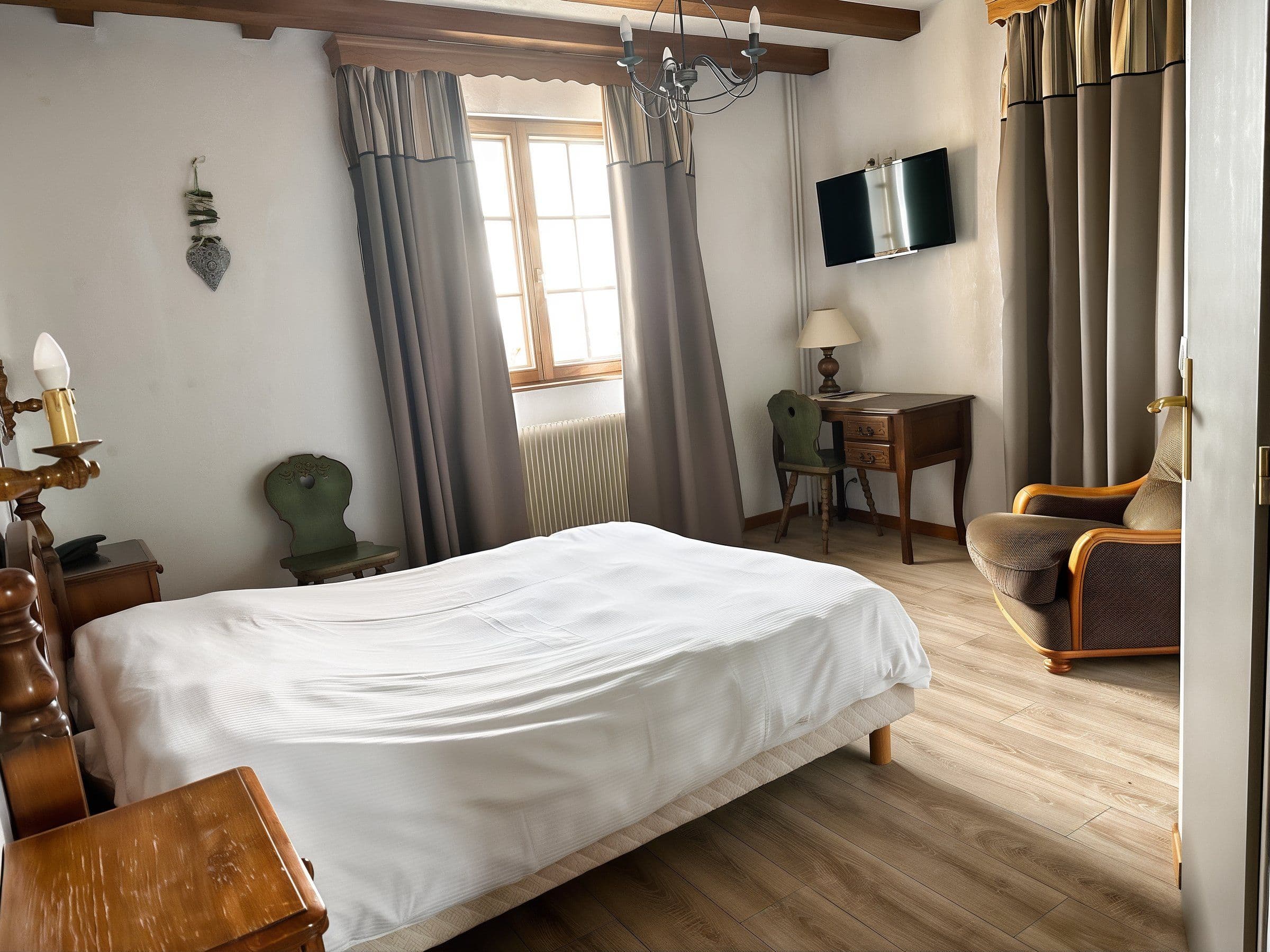 Chambre double lumineuse au style traditionnel alsacien avec poutres apparentes, grand lit en linge blanc, bureau en bois et fauteuil confortable près de la fenêtre.