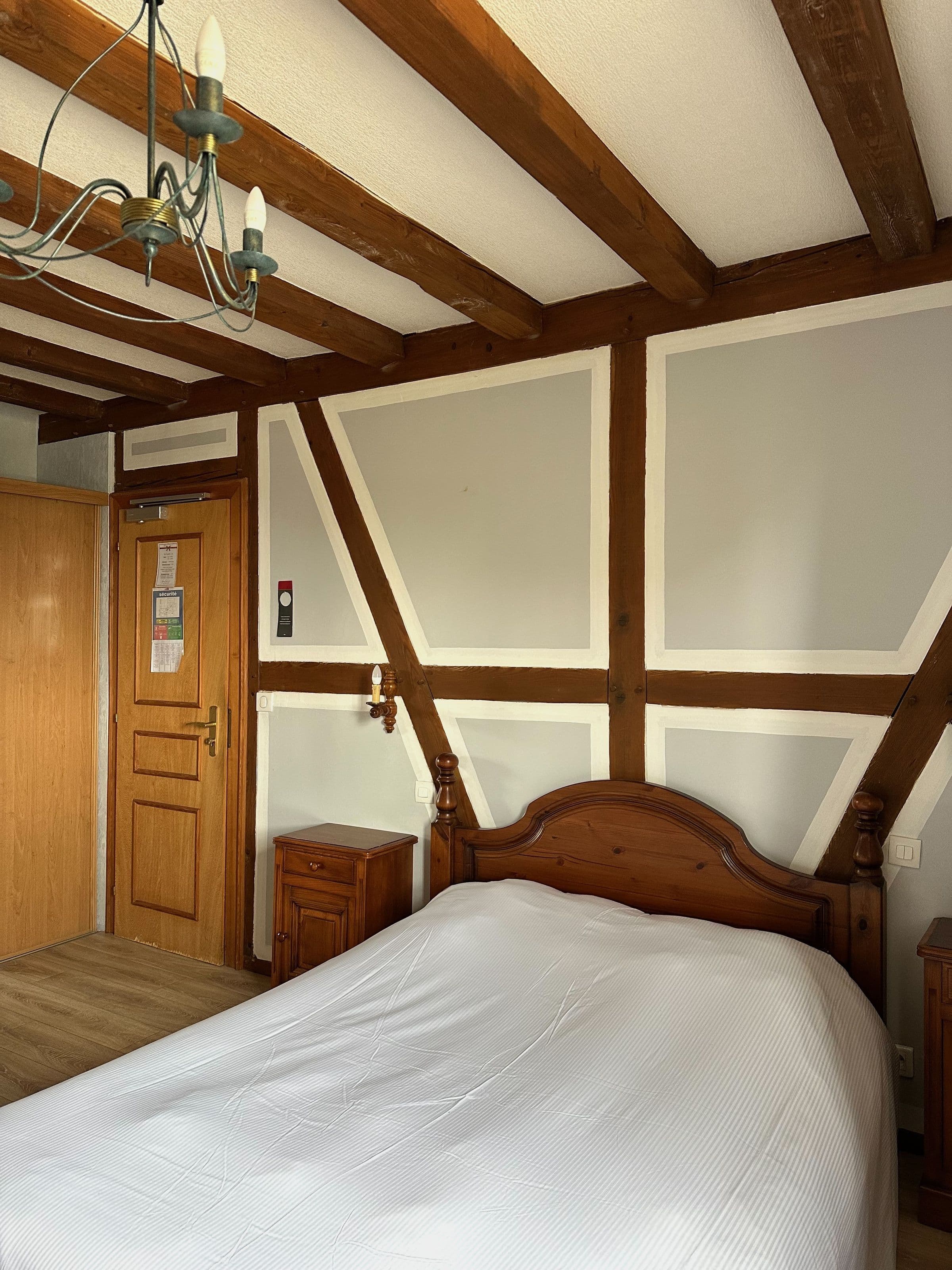 Chambre double chaleureuse de style alsacien avec poutres apparentes, mur à colombages, lit avec draps blancs et mobilier en bois.
