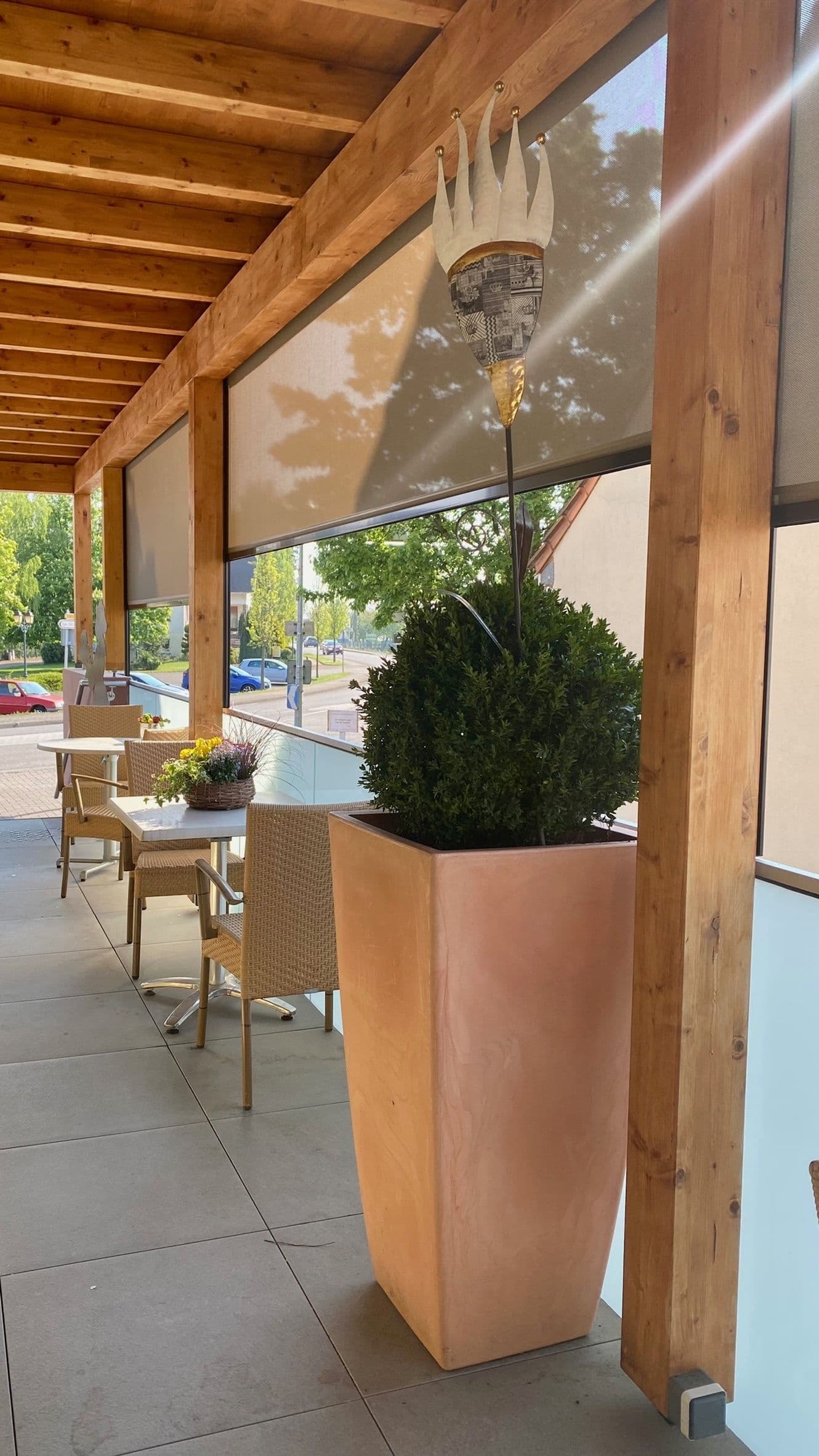 Terrasse couverte ensoleillée avec poutres en bois, chaises tressées, petites tables et grands pots en terre cuite contenant des buis, idéale pour déjeuner en plein air.