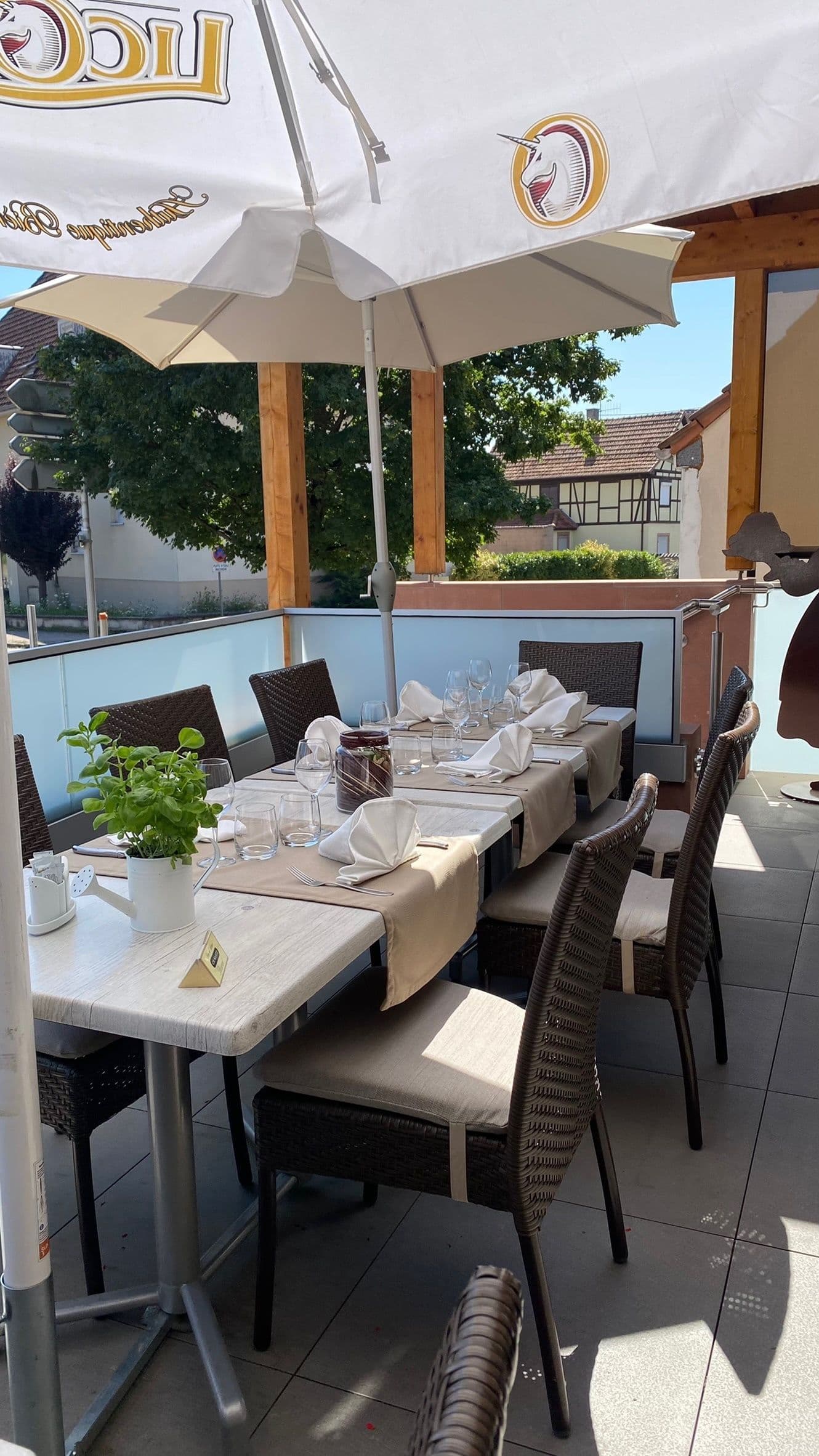 Terrasse ensoleillée du restaurant avec tables dressées, serviettes pliées, verres et une plante aromatique en pot sous de grands parasols et une pergola en bois, vue sur des maisons à colombages.