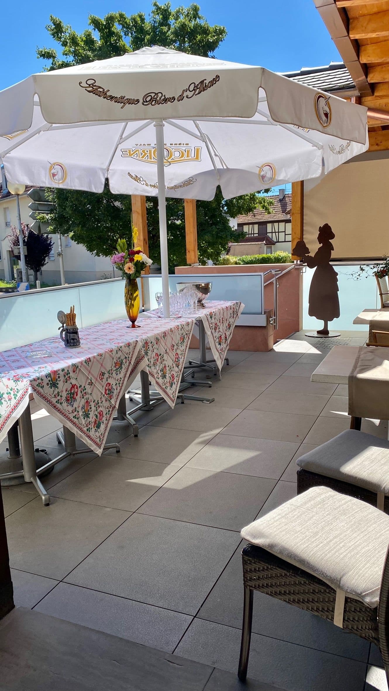 Terrasse extérieure ensoleillée de l'hôtel avec longues tables nappées de motifs floraux, grand parasol marqué, verre et vase de fleurs, prête pour un accueil convivial.