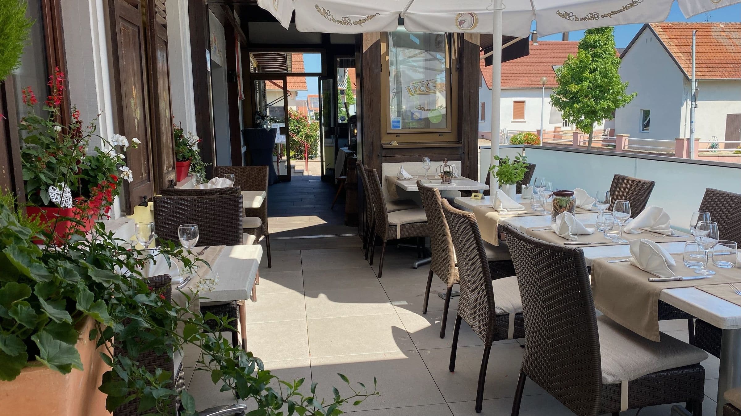 Terrasse ensoleillée du restaurant avec tables dressées, chaises en osier et parasols, entourée de fleurs et de végétation près du bâtiment à colombages.