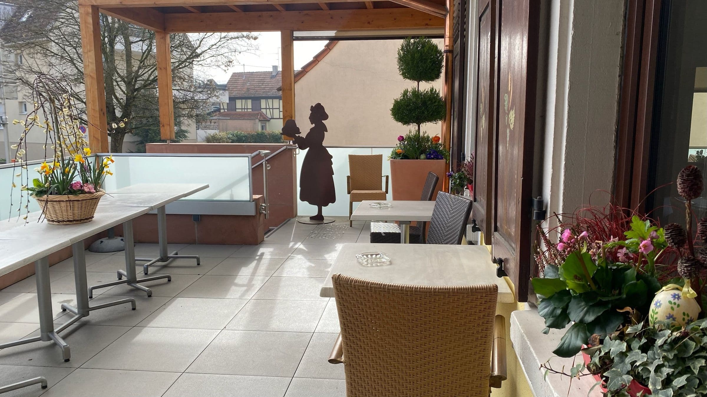 Terrasse couverte ensoleillée de l'Hôtel-Restaurant À La Couronne avec tables et chaises en osier, paniers de fleurs et topiaire, et une silhouette décorative en métal rappelant le charme alsacien.