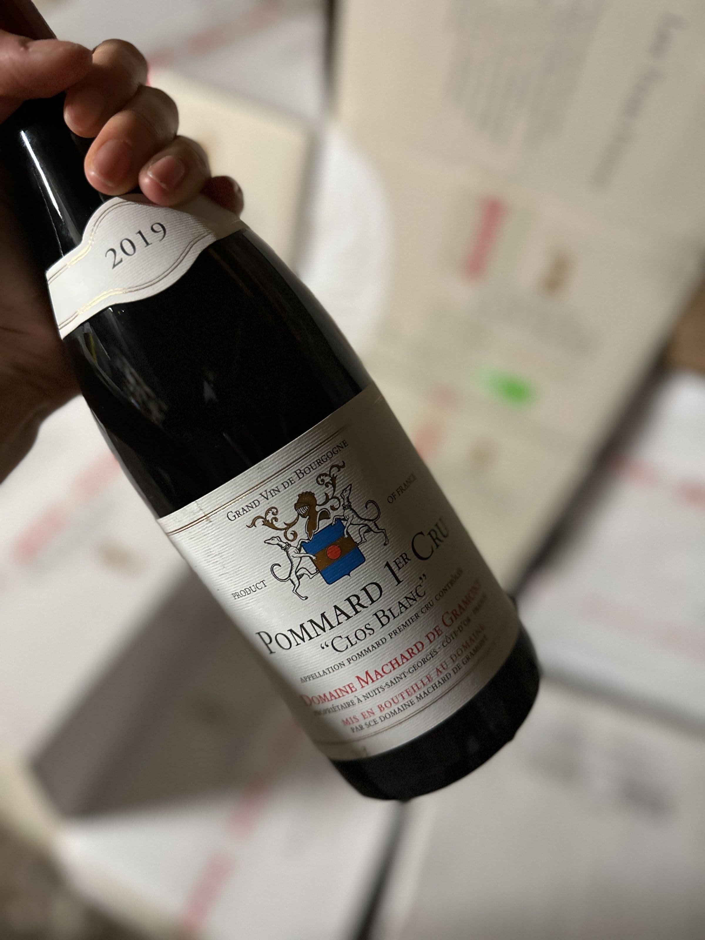 Gros plan d'une main tenant une bouteille de Pommard 1er Cru « Clos Blanc » 2019 du Domaine Machard de Gramont; l'étiquette et le millésime sont lisibles, l'arrière-plan est flou.