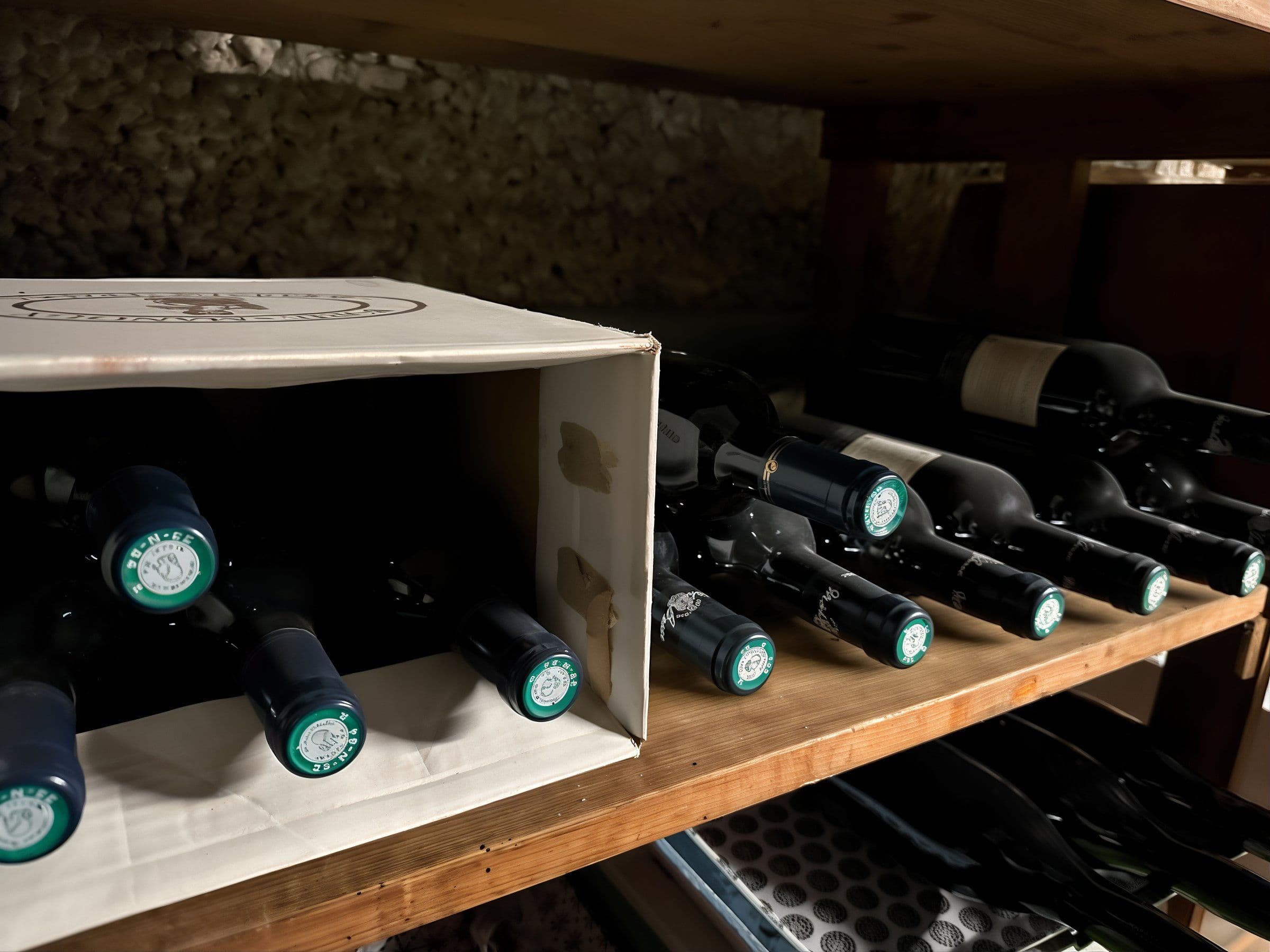 Gros plan sur des bouteilles de vin couchées sur des clayettes en bois et partiellement rangées dans une caisse en carton, avec des capsules vertes visibles.